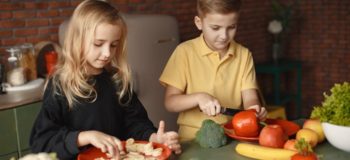 Focused little girl and boy cutting various vegetables and fruits while sitting at table and cooking in loft kitchen with brick wall