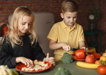 Focused little girl and boy cutting various vegetables and fruits while sitting at table and cooking in loft kitchen with brick wall